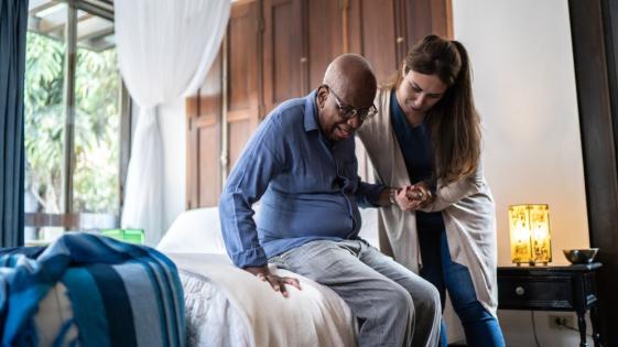 an older man sitting on a bed being helped onto his feet by a woman